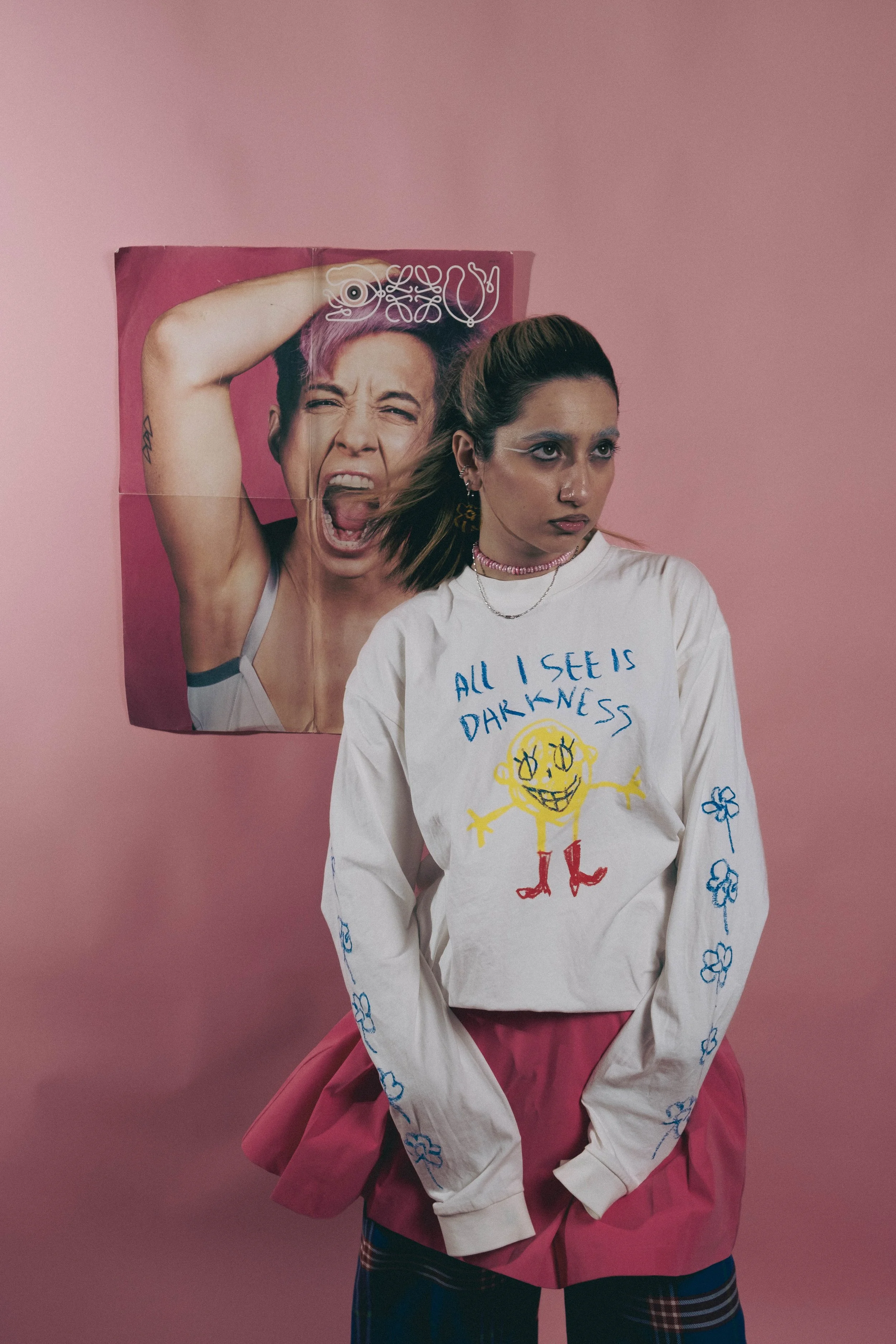 Person standing in front of a poster on a pink background, wearing a white shirt with 'All I see is darkness' and cartoon design, pink skirt, and plaid pants.