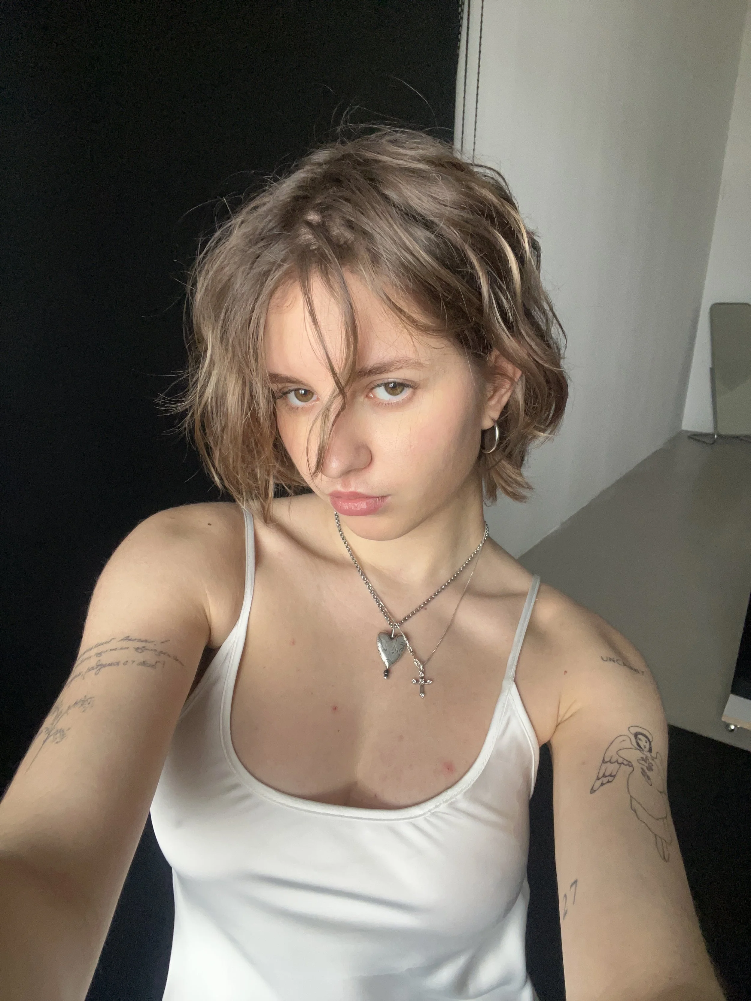 Person with short wavy hair wearing a white tank top and layered necklaces. Visible tattoos on both arms.