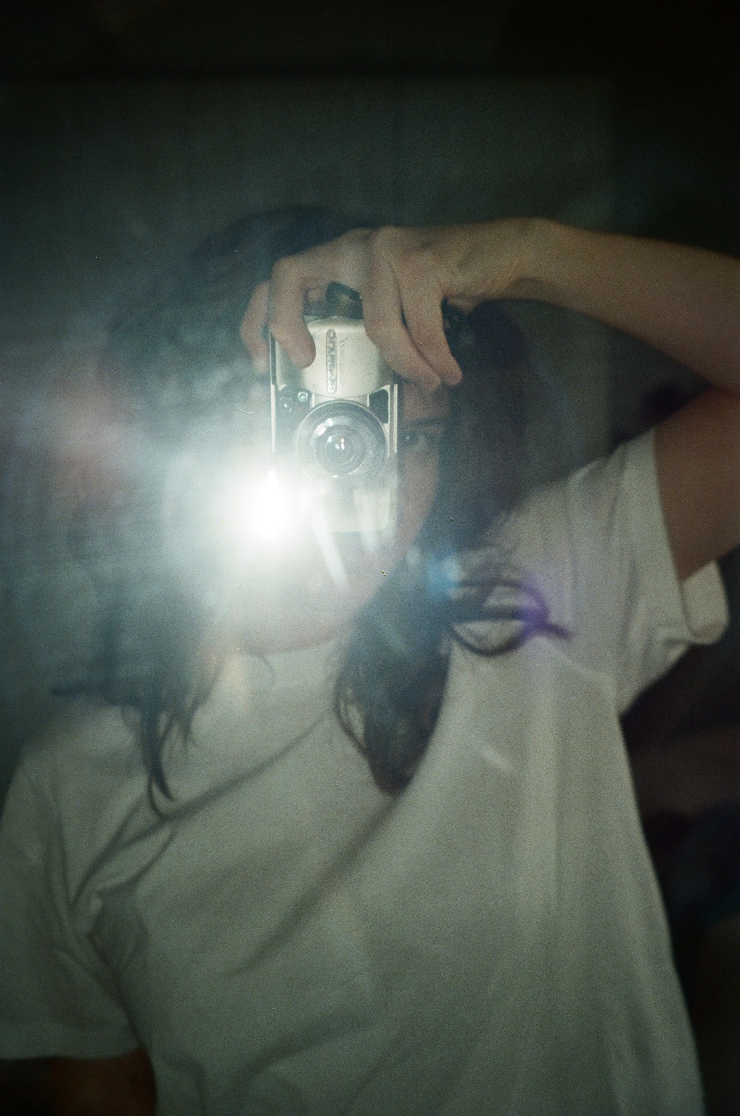 Person taking a photo with a vintage camera, wearing a white shirt, flash reflecting in a mirror.