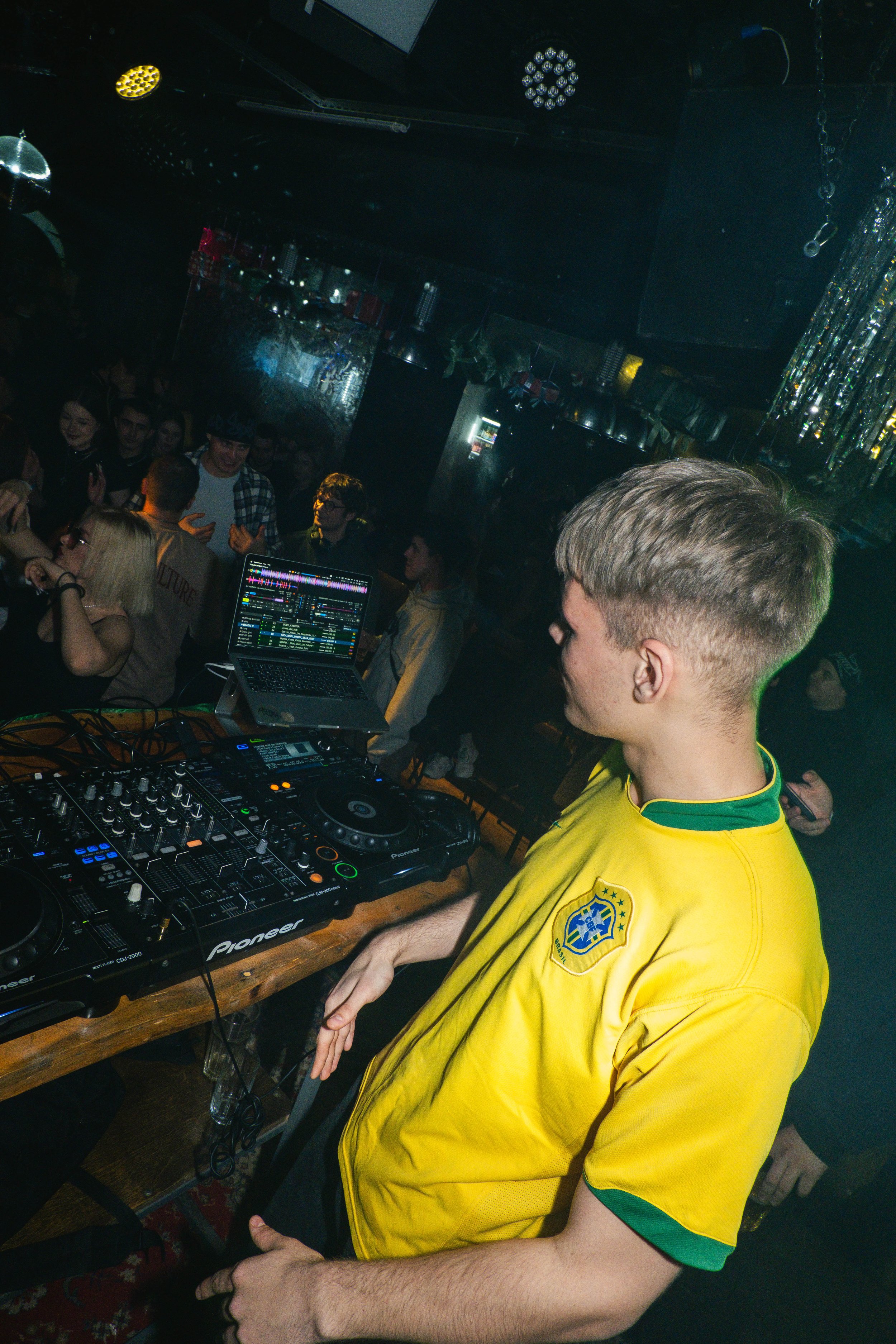 DJ in a yellow shirt playing music at a nightclub with a crowd in the background.