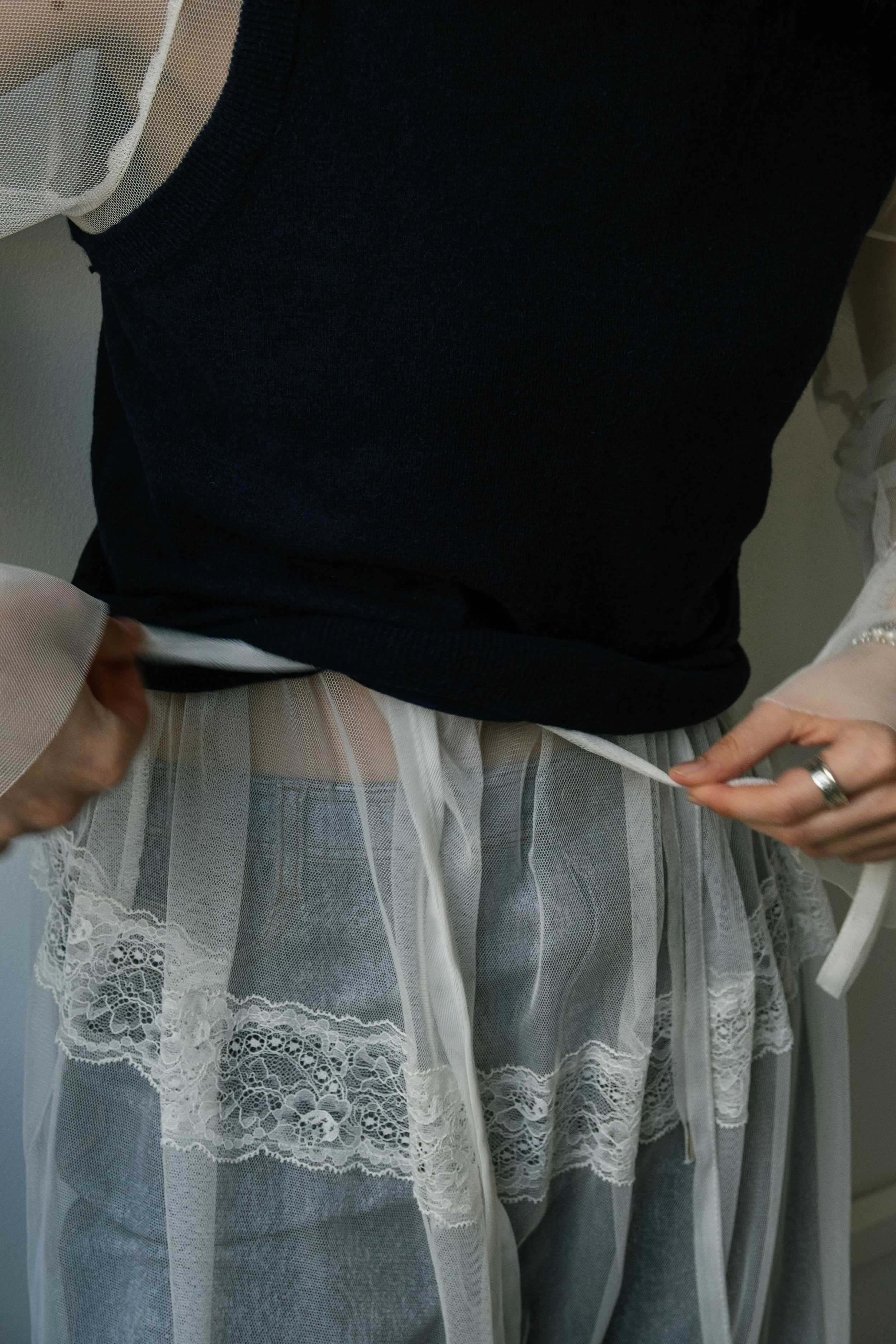 Person wearing a black top over a sheer, lace-trimmed layer and gray jeans, tying a white ribbon around their waist.