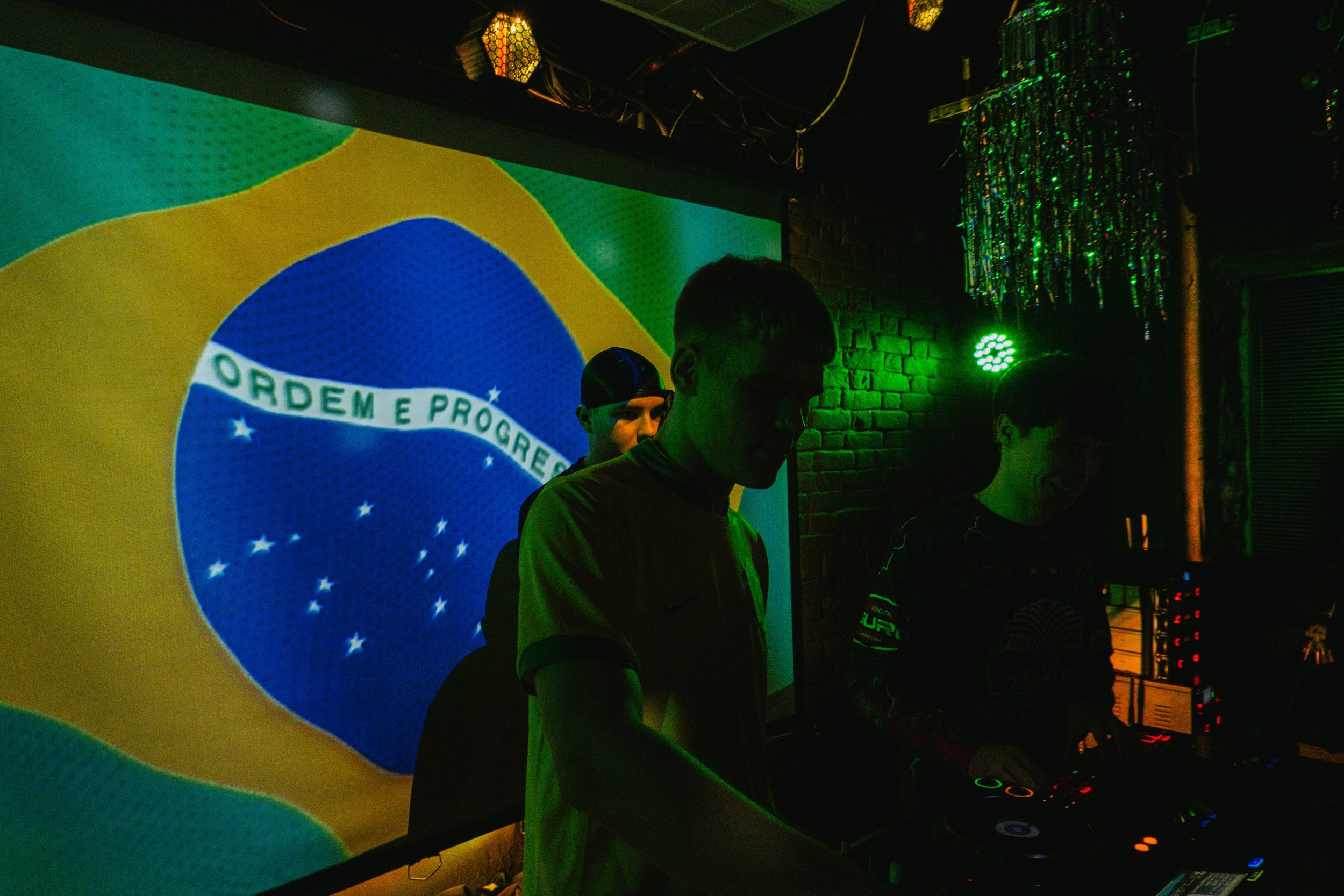 People DJing in a dimly lit room with the Brazilian flag projected on a screen behind them.