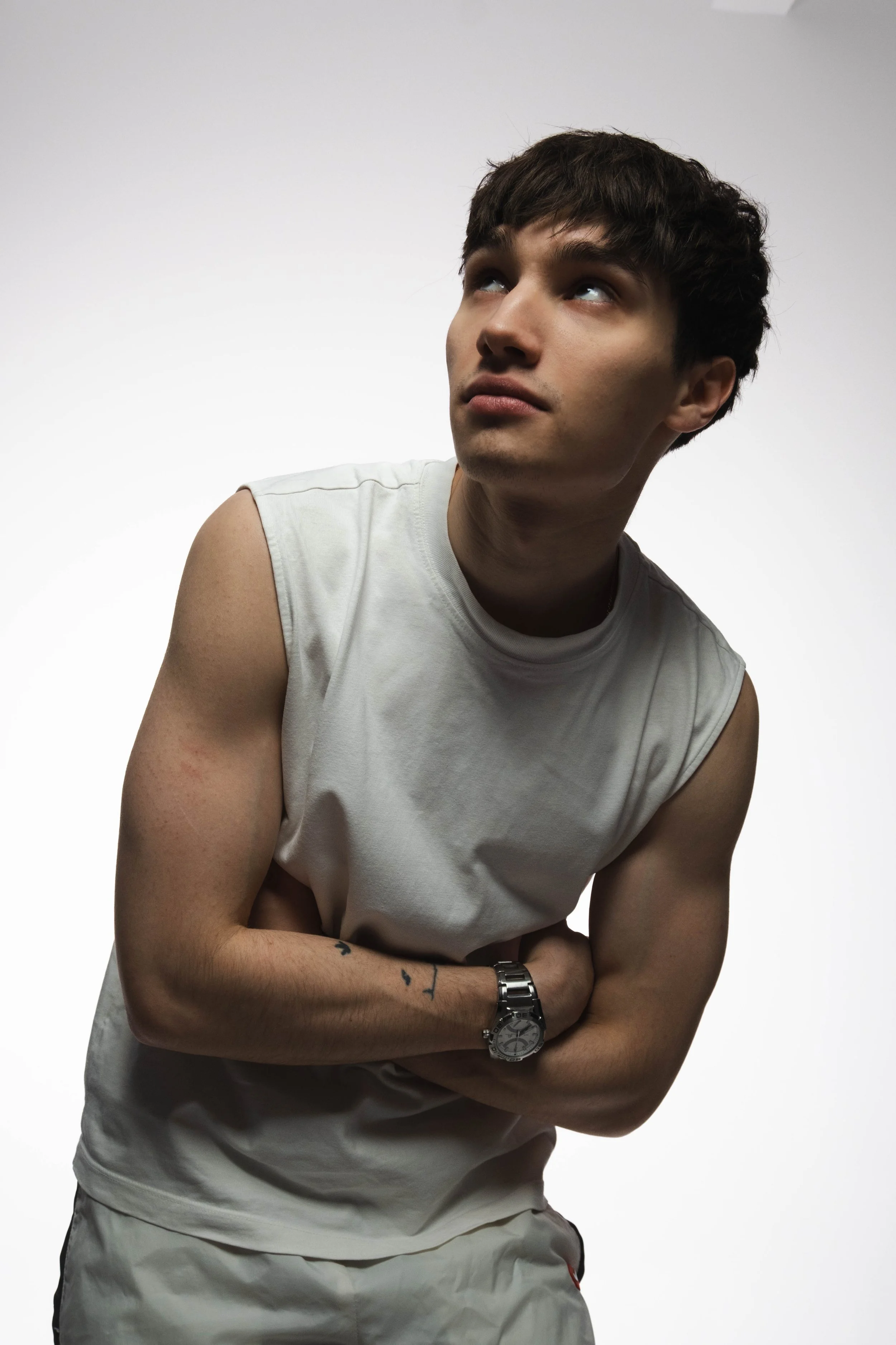 Young man in a white sleeveless shirt and watch, looking upwards against a plain background.