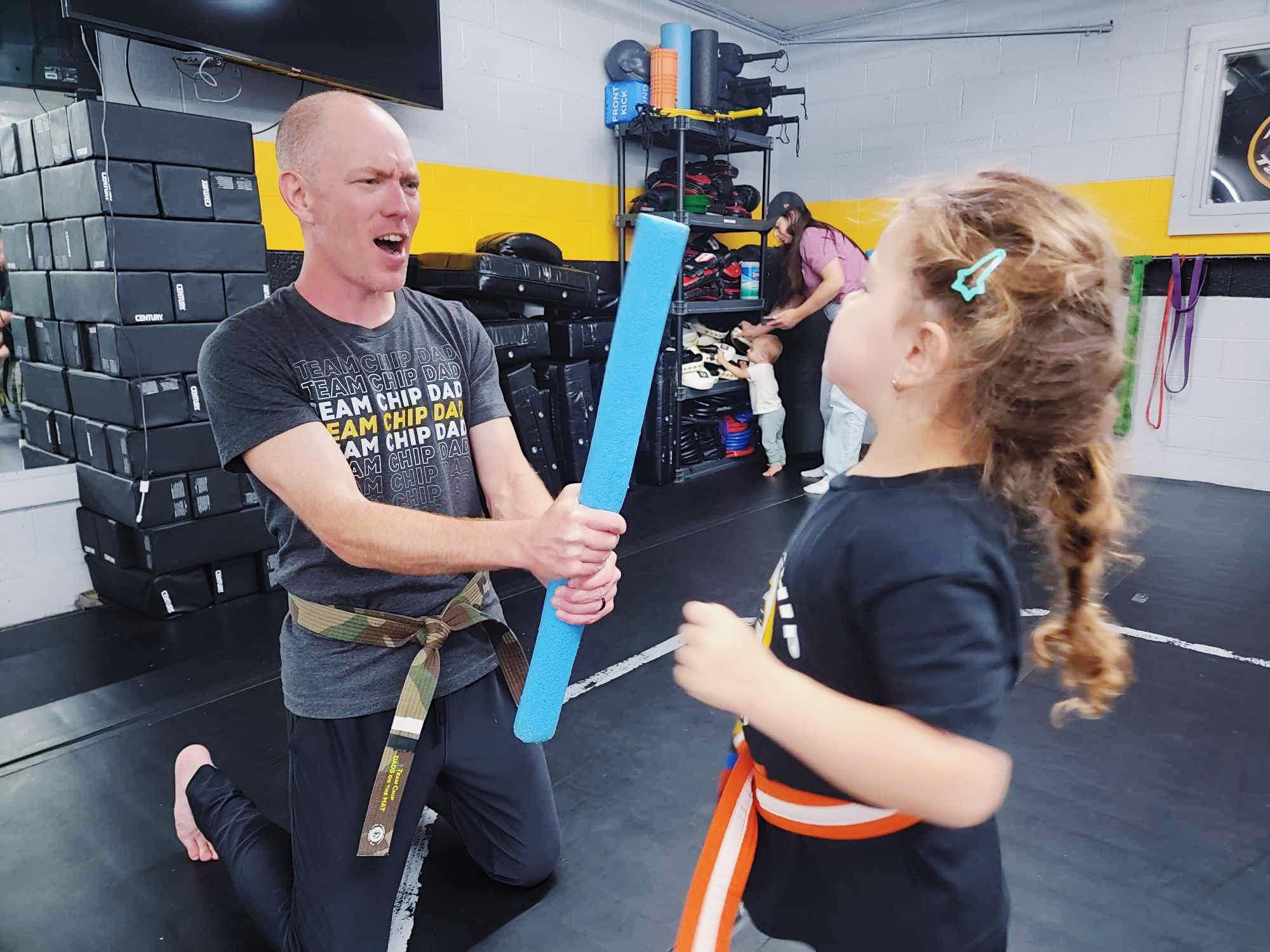 Martial arts class with instructor holding foam stick and child with orange belt, inside gym with equipment in background.