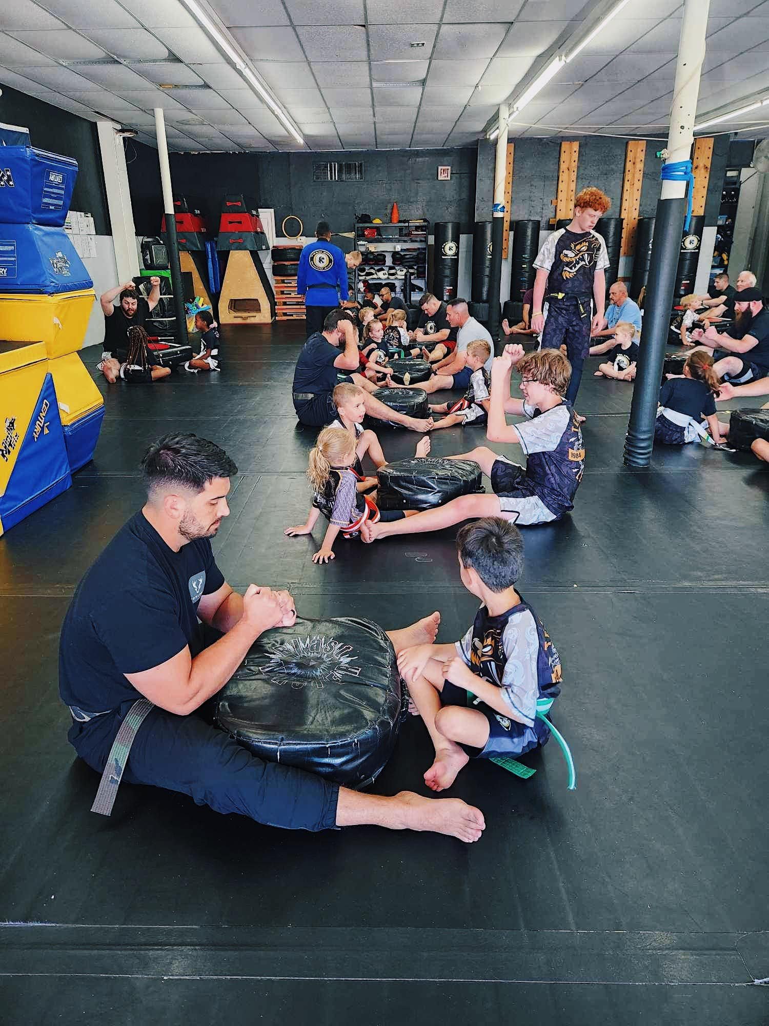 A group martial arts class with adults and children practicing on a mat. Participants are sitting on the floor holding pads or engaging in exercises. The room is equipped with martial arts gear and equipment along the walls.
