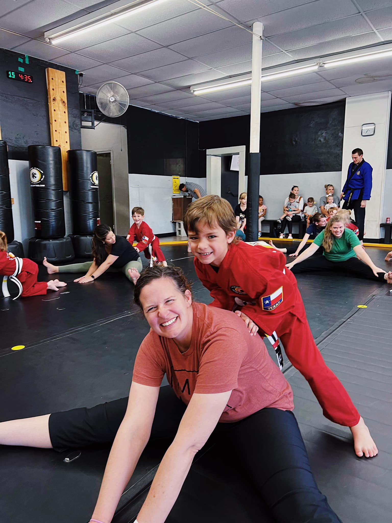 A group of people, including children and adults, practicing martial arts stretches on a mat in a training room. Some individuals are wearing martial arts uniforms, and others are in casual athletic clothing. The environment appears lively and engagi