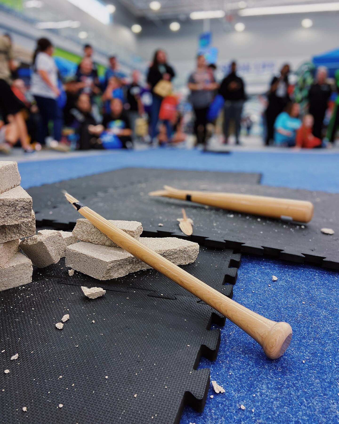 Broken wooden bats on gym mats with shattered cinder blocks, surrounded by a crowd in a gymnasium.