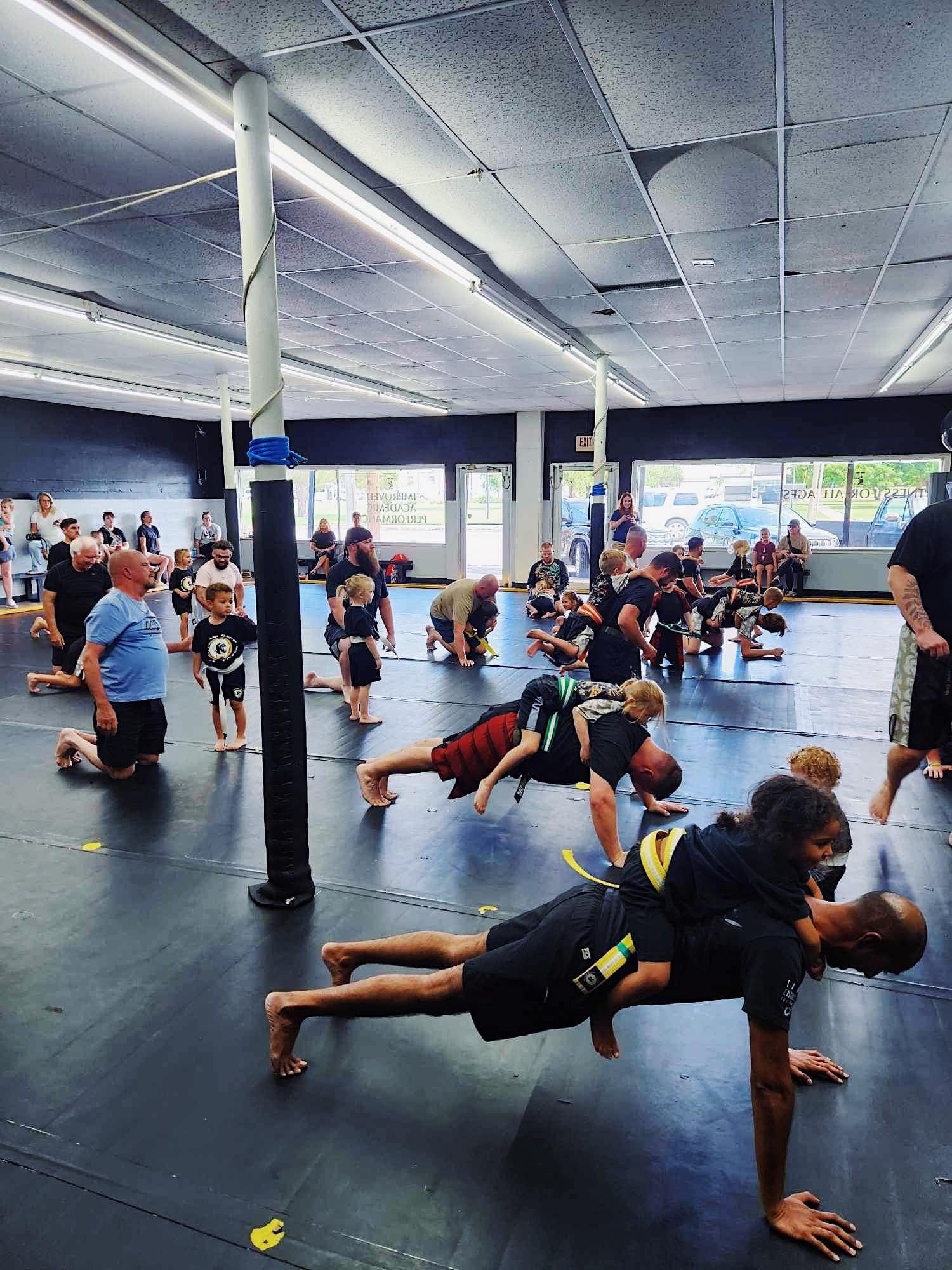 Children and adults practicing martial arts exercises on mats in a gym, some adults doing push-ups with kids on their backs. Spectators watching from the sides, windows in the background.