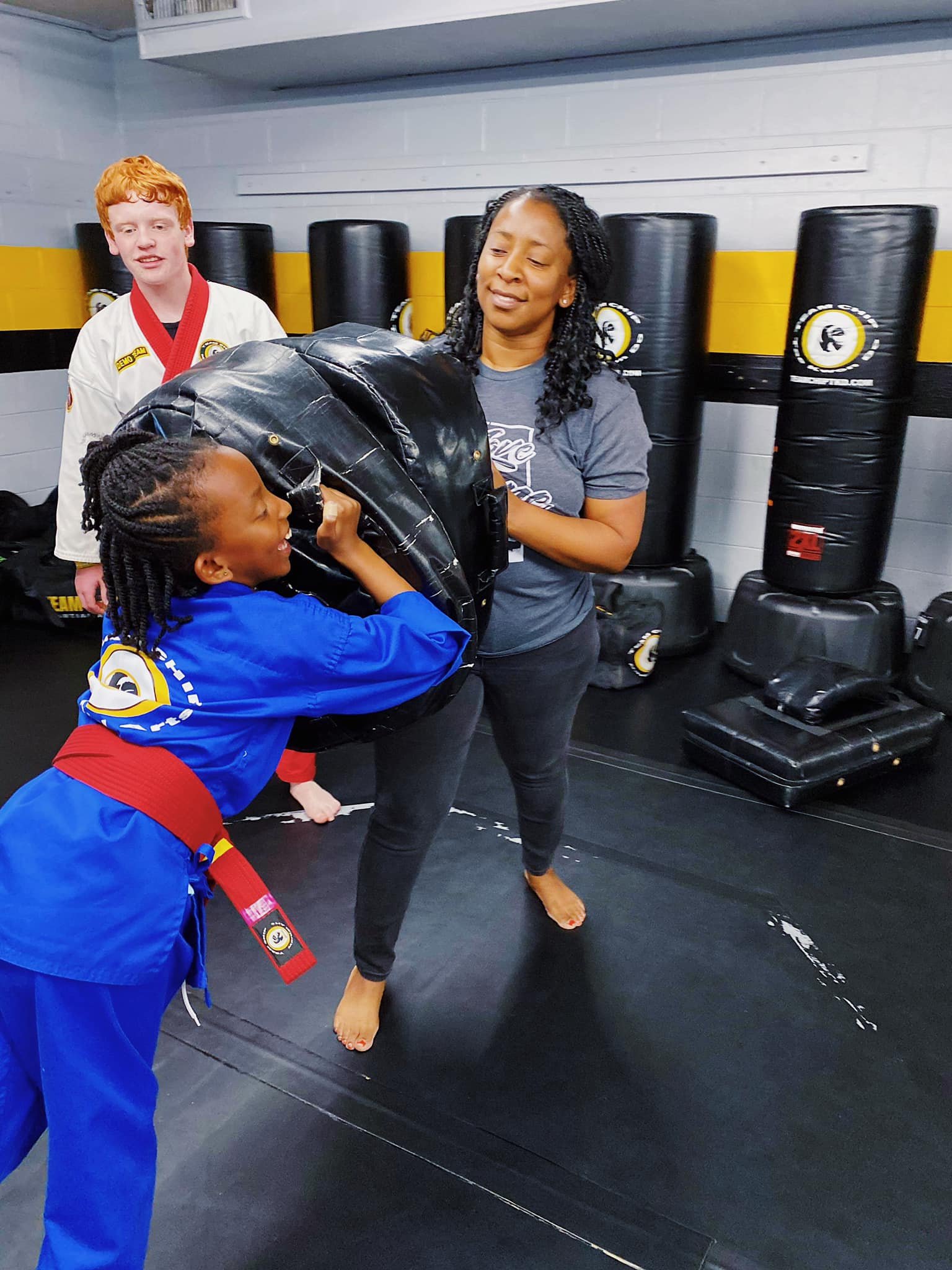 A young martial arts student practicing a move on a padded shield held by a woman in a training room, with another student in a gi observing in the background.