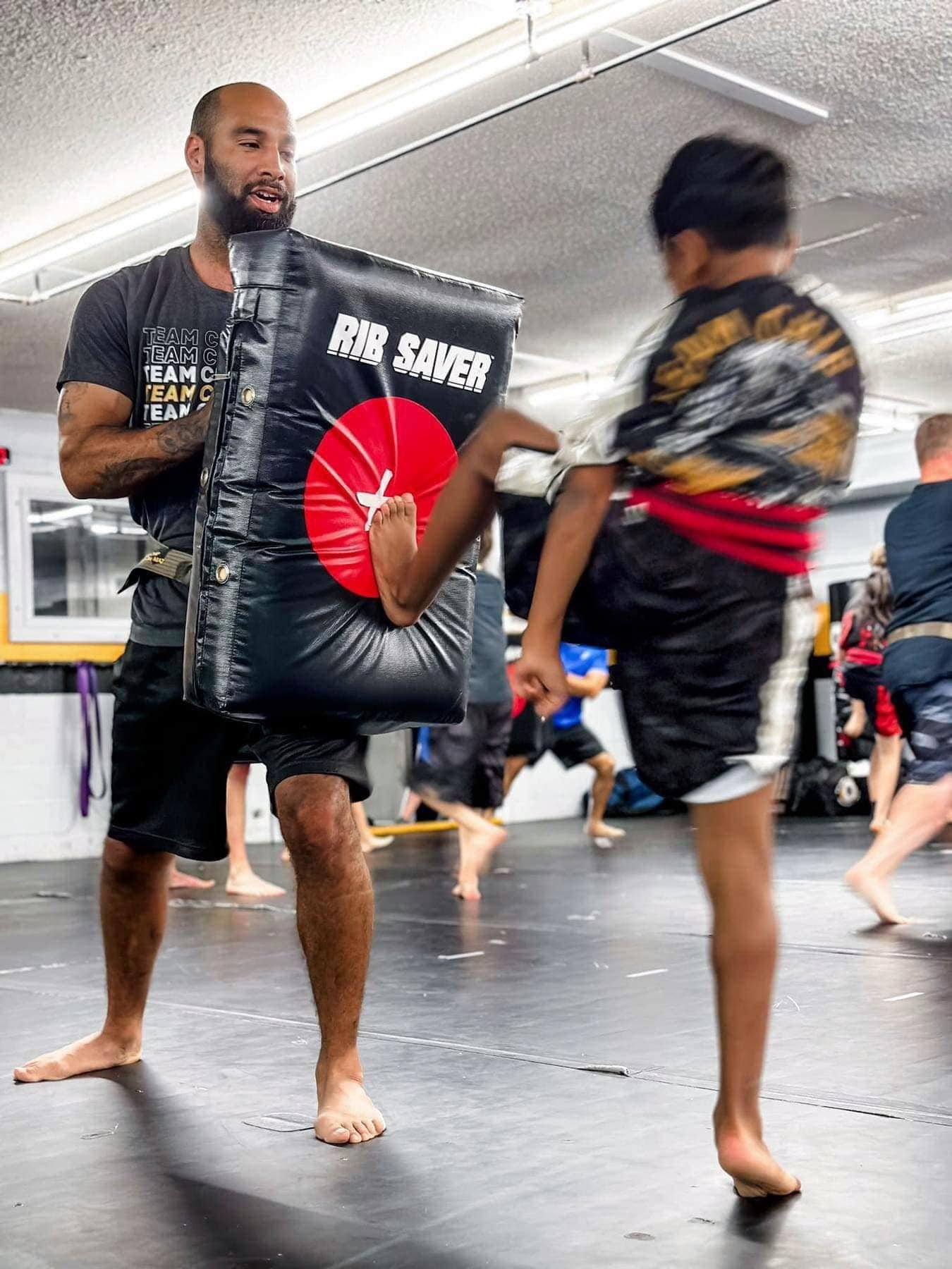 A martial arts class in session with a man holding a kick shield labeled "Rib Saver," while a child executes a kick towards it in a gym setting.