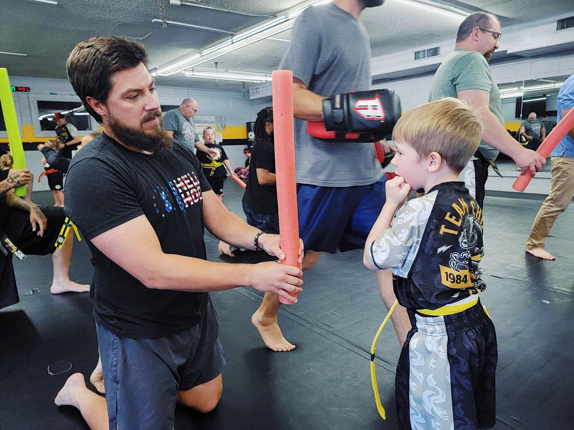 Martial arts class with instructor and child holding padded sticks, wearing uniforms and yellow belts.