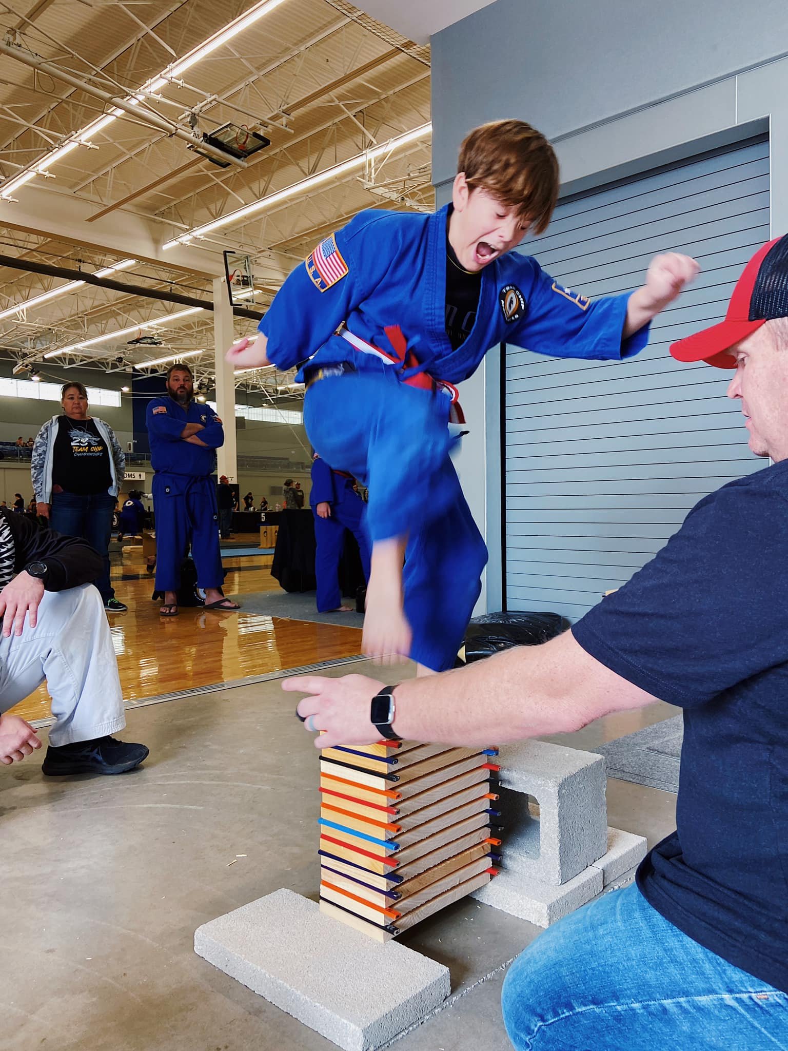 A young martial artist in a blue gi breaking wooden boards with a jump kick at a martial arts event. Several people are watching, including a person holding the boards in place. The environment appears to be a sports or community center with an indoo