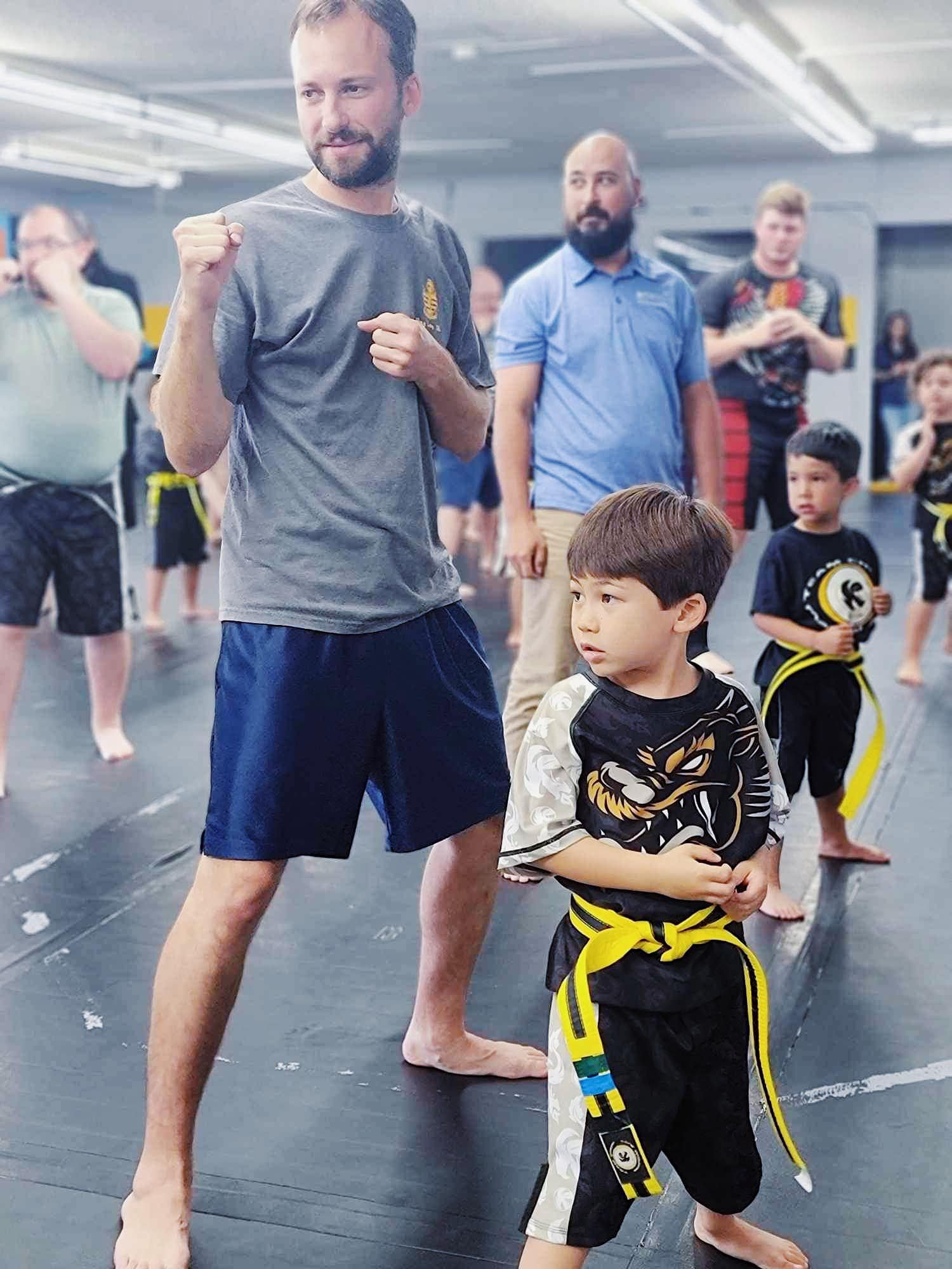 Adults and children practicing martial arts in a gym, with a focus on a man and a boy standing in a fighting stance on a mat. The boy has a yellow belt.