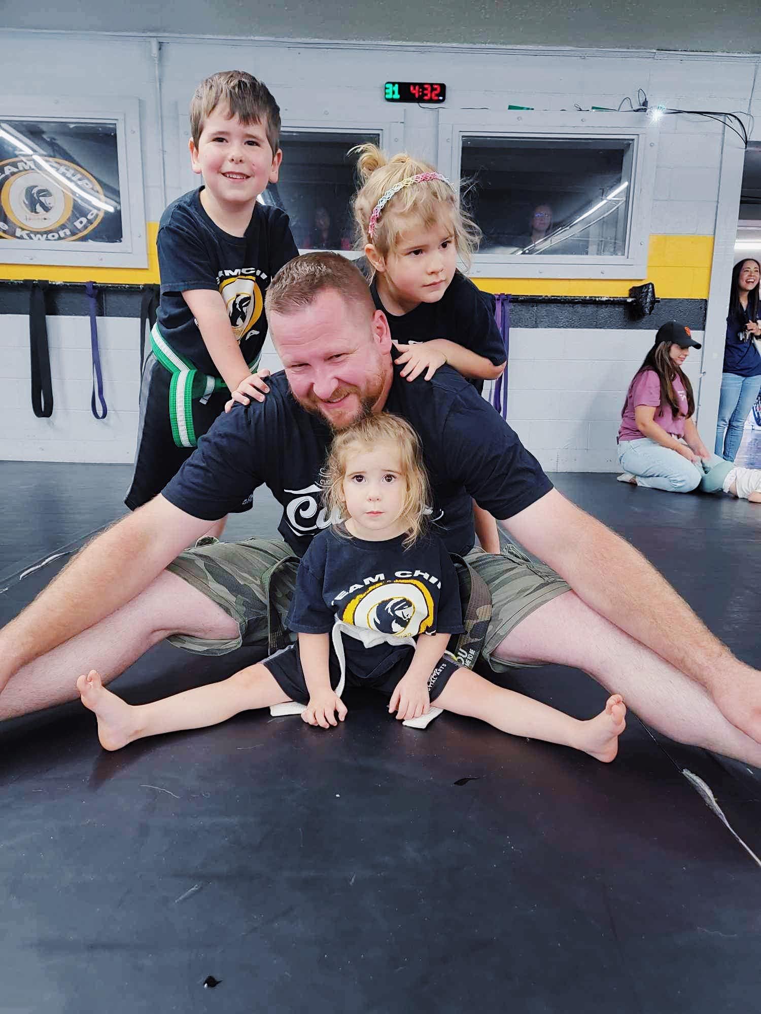 A man sitting on a martial arts mat with three young children. The man is spreading his legs in a stretching pose, and the children are positioned around him. One child is sitting in front, and the other two are standing behind him, smiling. They are