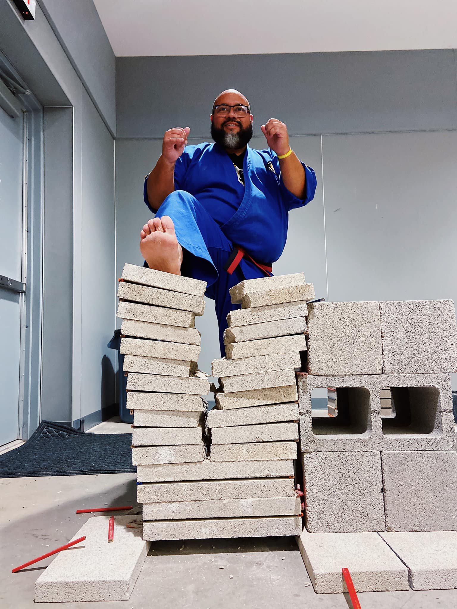 Man in blue karate uniform breaking concrete blocks with a kick inside a room.