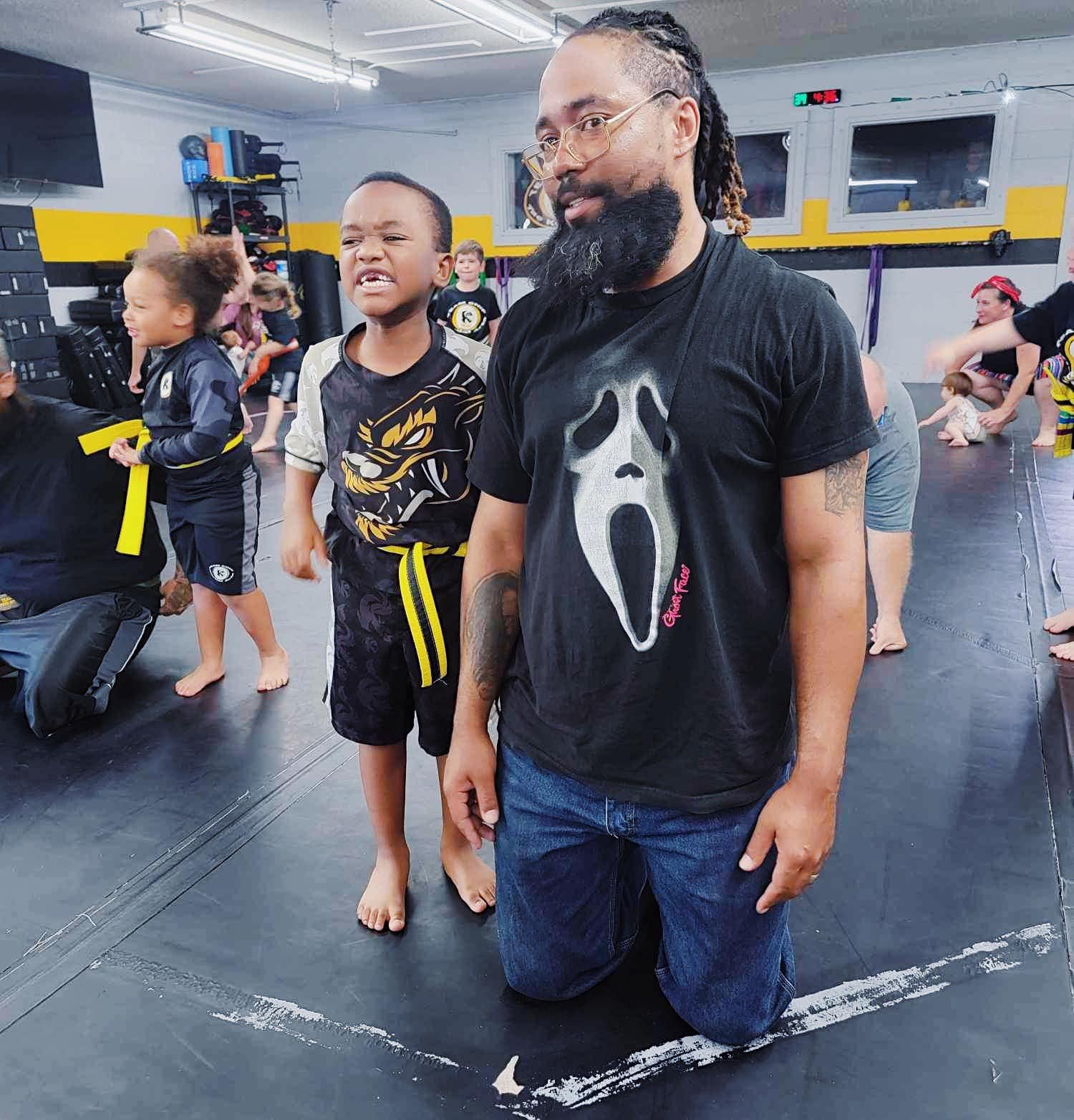 Children in a martial arts class with an adult instructor kneeling. One child is wearing a black and yellow uniform, while the instructor is wearing a t-shirt with a ghost face design.