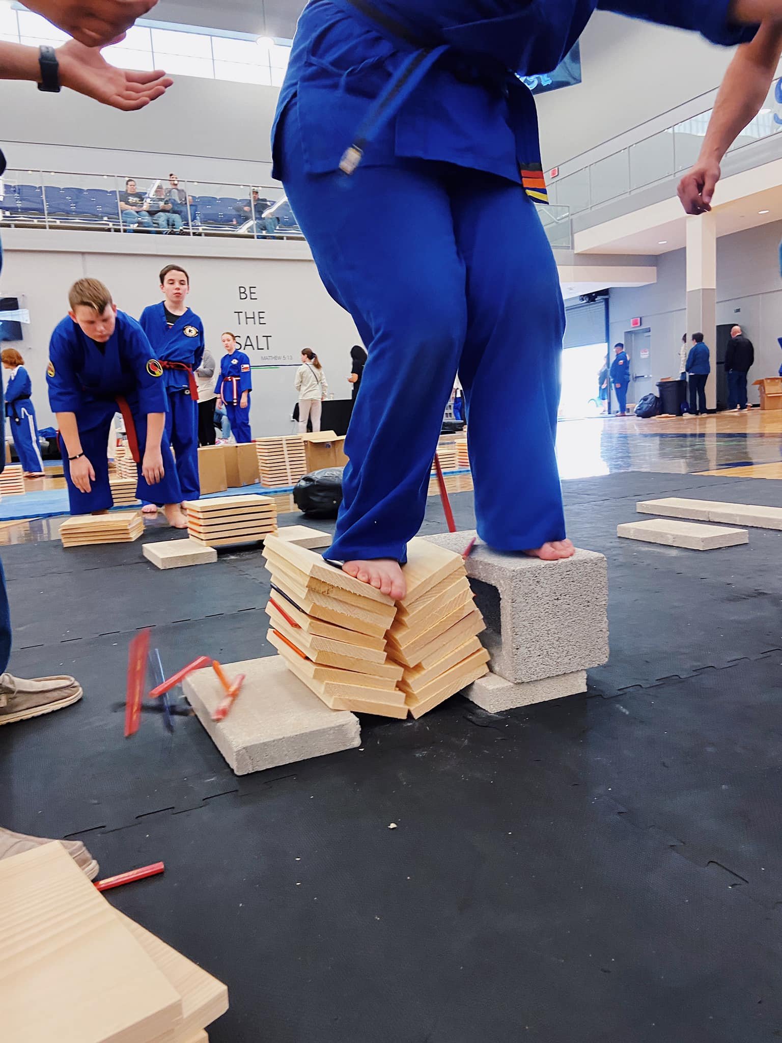 Martial arts practitioner in blue uniform breaking wooden boards with foot at a dojo event, surrounded by spectators and other participants.
