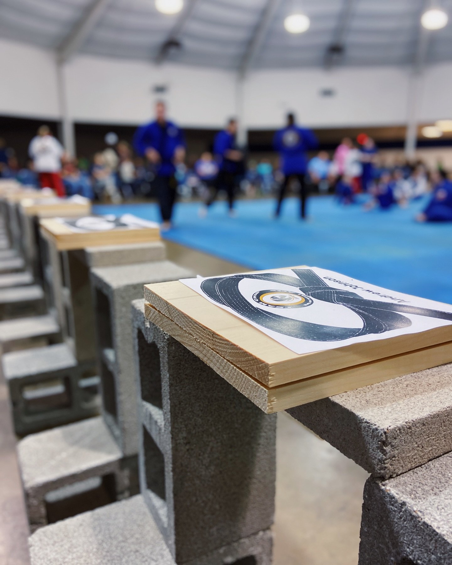 Cinder blocks and wooden boards set up for a martial arts demonstration in a gymnasium. People in martial arts attire are in the background.
