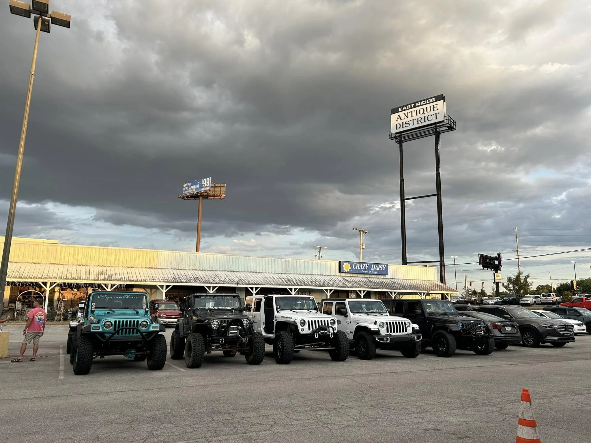 Parking lot with six Jeep vehicles parked in front of a restaurant named Crazy Daisy, under a cloudy sky, with billboards overhead and a lone person walking past