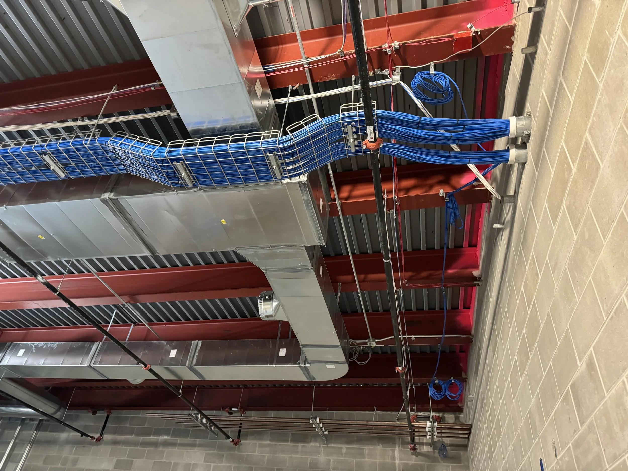 Industrial mechanical room with red steel beams, metal ductwork running along the ceiling, and blue electrical cables secured with cable ties.
