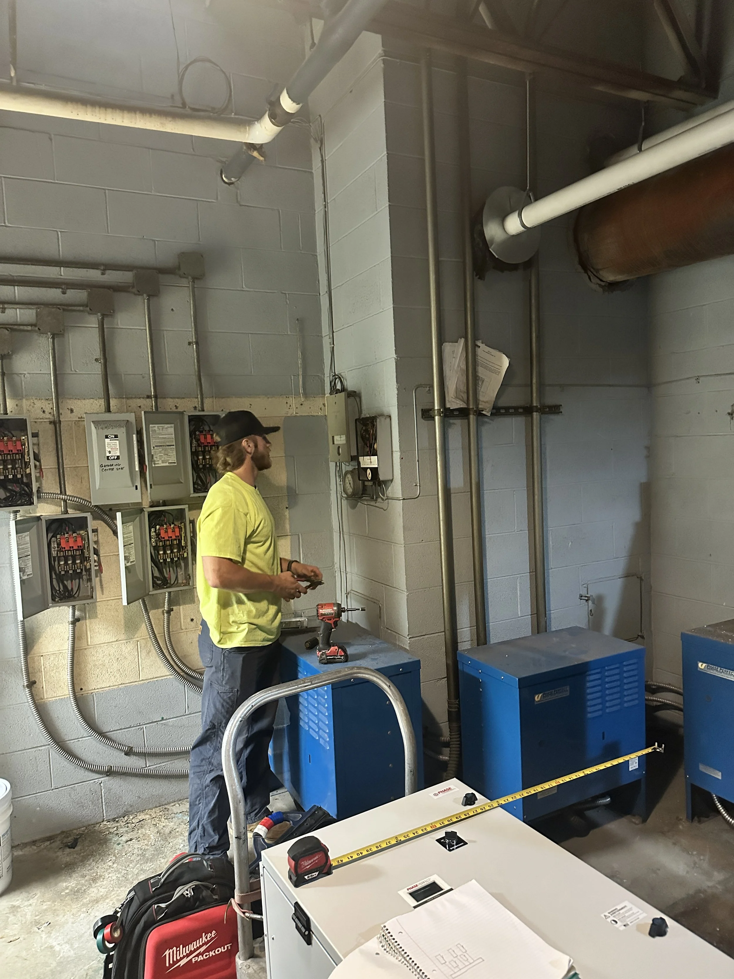 A man in a yellow shirt and black cap working in an industrial or utility room with electrical panels, pipes, and equipment. There are tools and equipment on a table, including a tape measure, coffee mug, and notebooks.