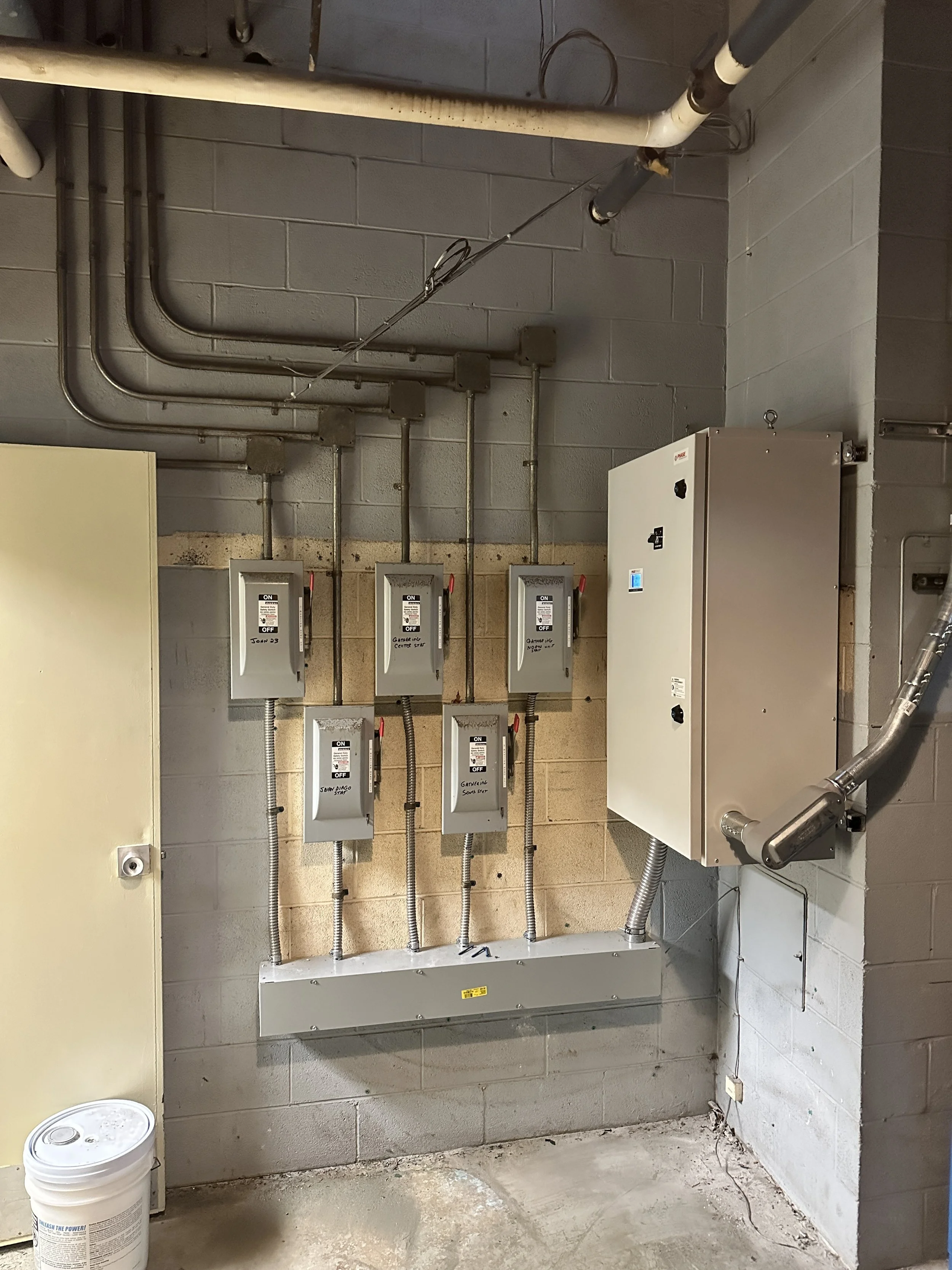 Electrical panel with six circuit breakers mounted on a concrete wall in an unfinished utility room, with pipes running along the wall and floor.