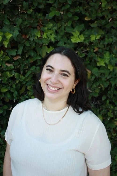 White woman with wavy dark brown hair, wearing a white top and smiling in front of green leafy background