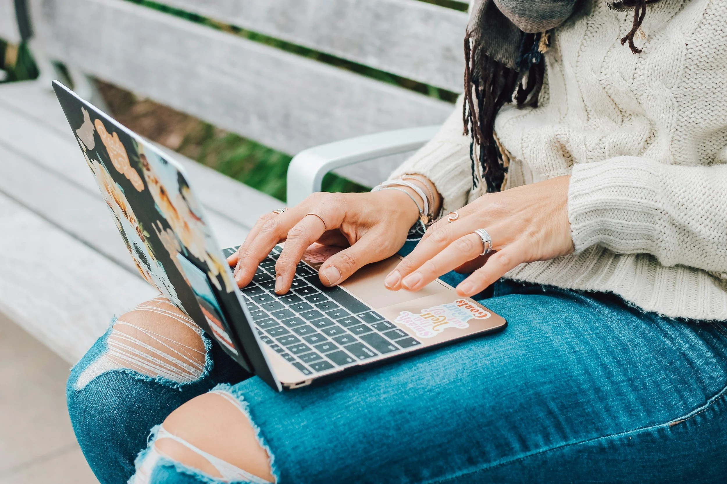 Person sitting on a bench using a laptop decorated with stickers, wearing ripped jeans and a cream sweater.