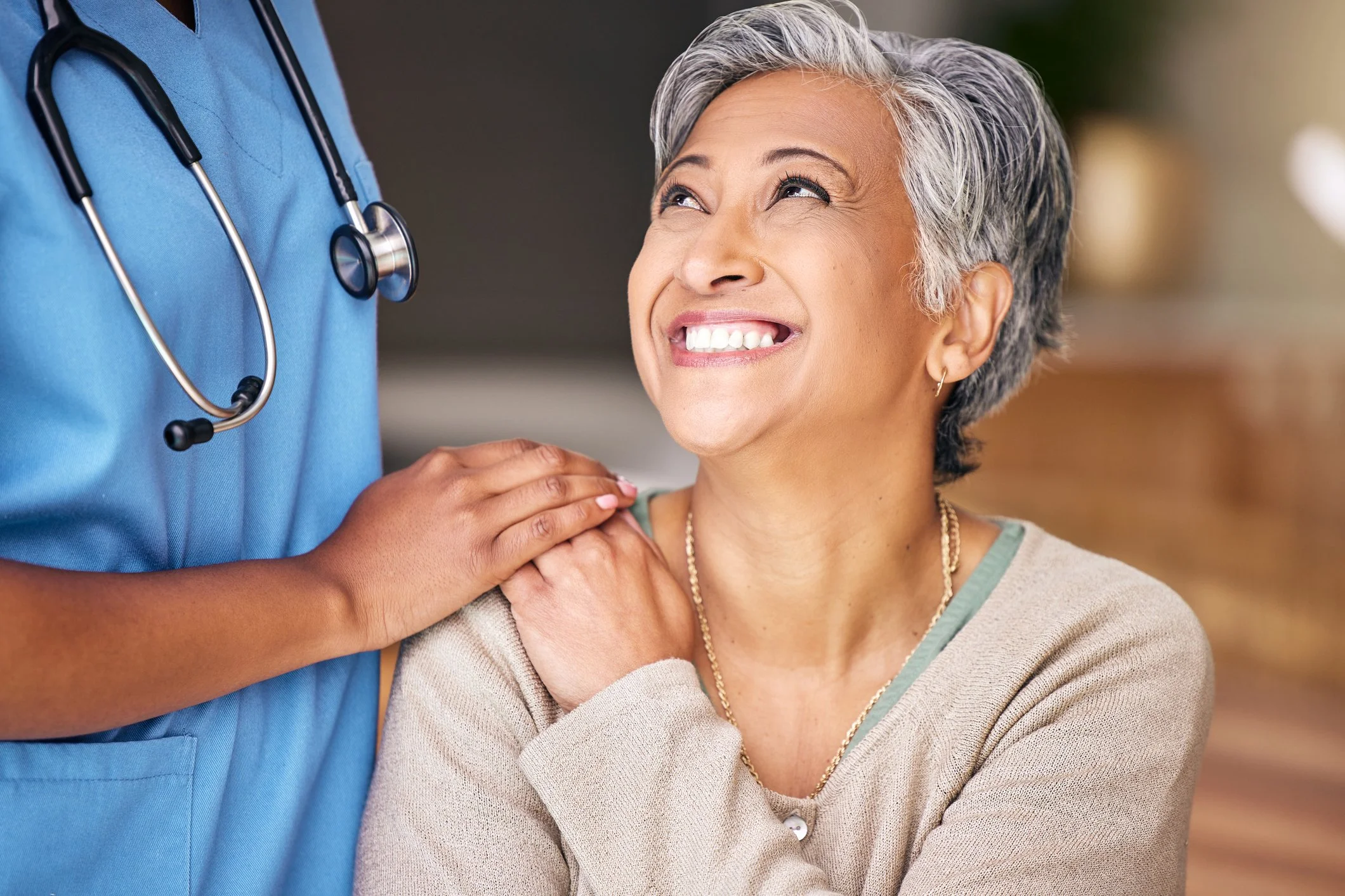 A smiling older woman with short gray hair looking up at a healthcare worker in blue scrubs. The healthcare worker has a stethoscope around their neck and is gently holding the woman's shoulder.