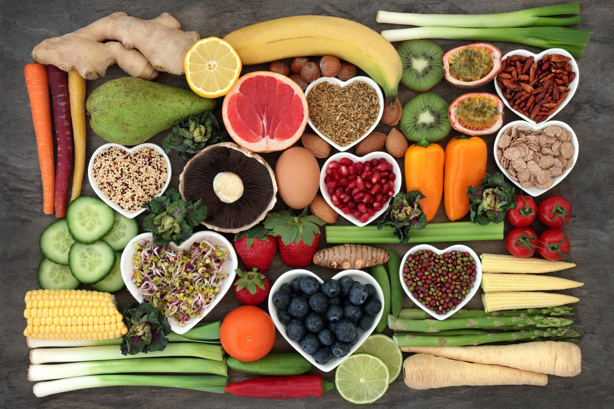 Assorted fresh fruits and vegetables arranged on a dark surface with some in heart-shaped bowls
