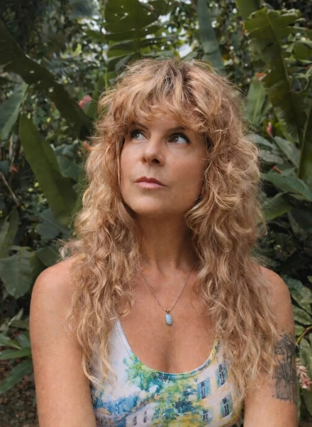 A woman with long, curly blonde hair posing outdoors among green foliage, wearing a colorful top and a blue pendant necklace.