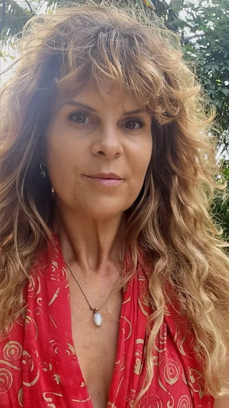 A woman with curly, light brown hair taking a selfie outdoors, wearing a red patterned top and a pendant necklace, with greenery in the background.