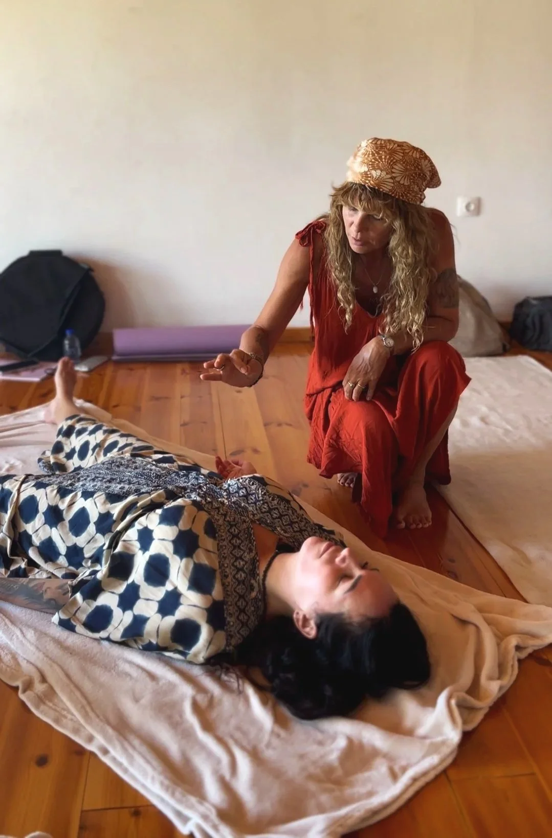 A woman in a red dress and a patterned hat kneeling beside a woman lying down on a bed with a blanket, holding her hand in a comforting manner.