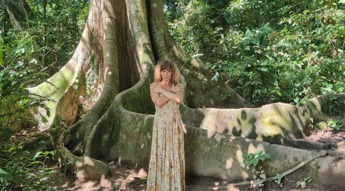 A woman with curly hair wearing a long, patterned dress stands with crossed arms in front of a large tree in a dense forest.
