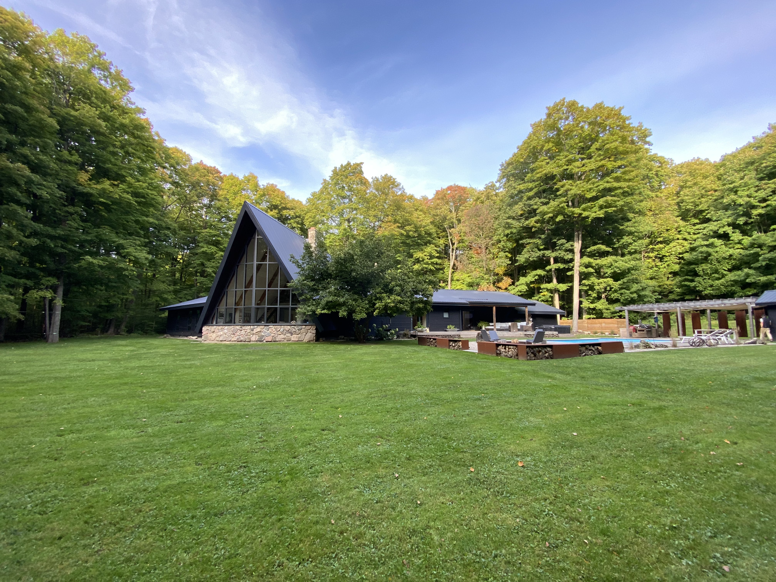 A-frame house with large windows and stone base surrounded by lush green lawn and trees, under a blue sky.