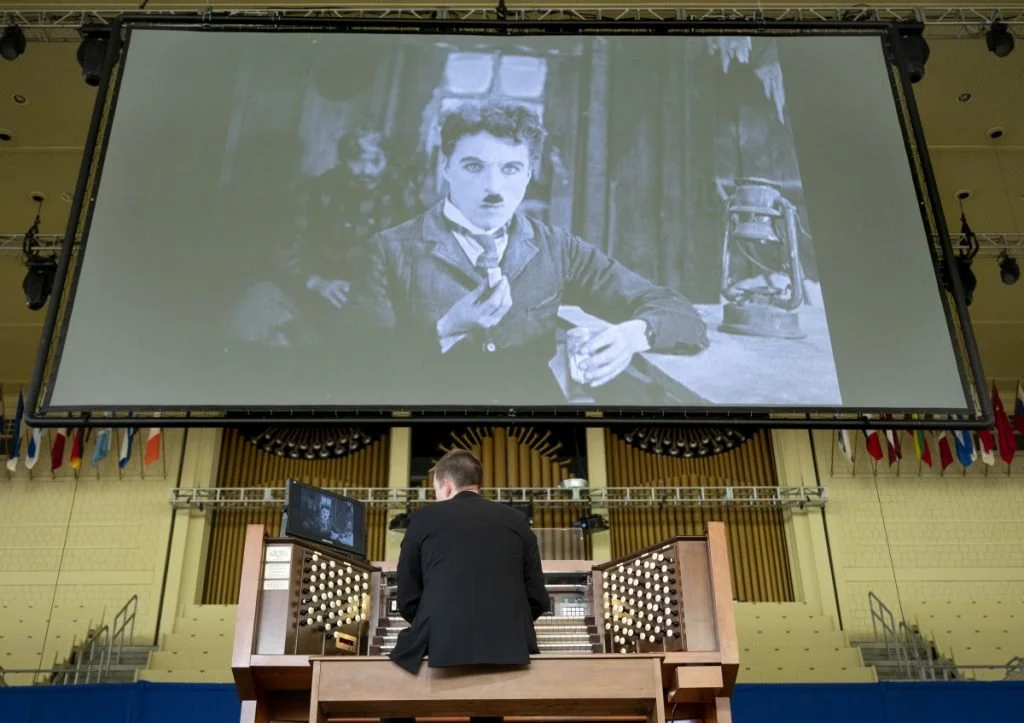 A person with short hair, dressed in a black suit, is playing a large pipe organ in a hall. In front of them, a large screen displays a black and white image of a woman in old-fashioned clothing, with short hair and a serious expression, sitting at a table with a glass and a lamp in the background.