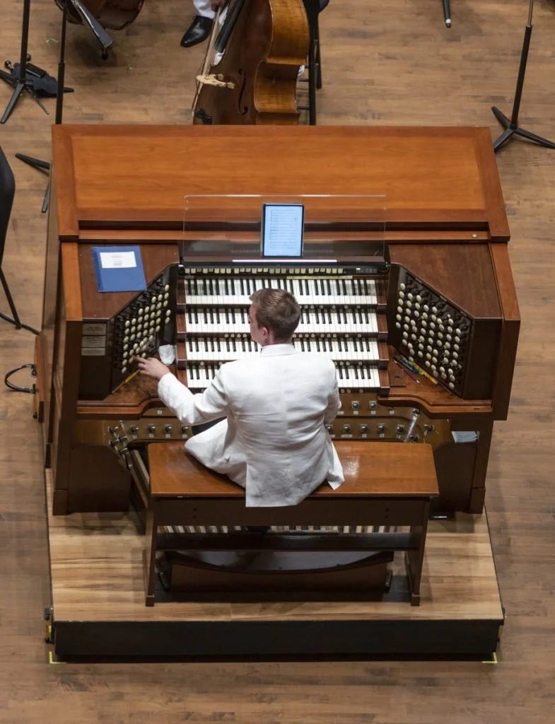 A person in a white coat playing a vintage church organ with multiple keyboards and stops, in a wooden-floored hall with other musical instruments and stands.