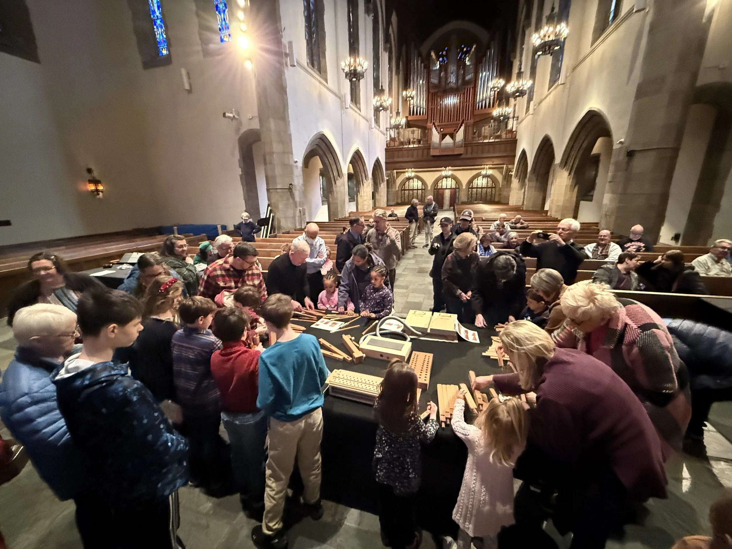 A large group of people gathering inside a church with high arched ceilings, stained glass windows, and a pipe organ at the back. Some individuals are seated on pews, while others are standing and engaging in activities or conversation.
