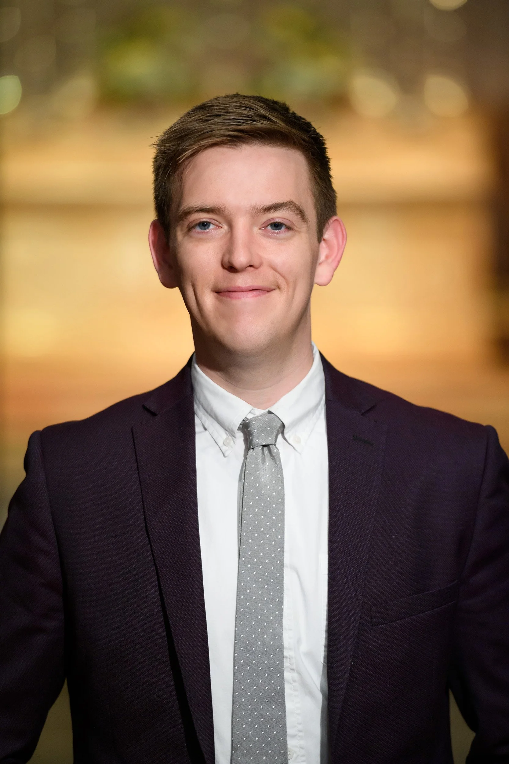 A young man with light skin, short brown hair, and blue eyes is smiling slightly. He is wearing a dark suit, white shirt, and light gray tie with small white polka dots. The background is blurred with warm colors.
