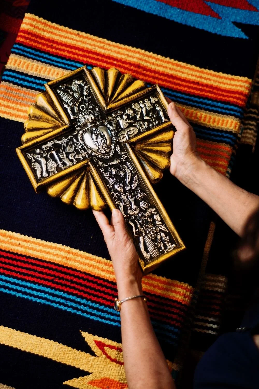 Person holding a silver and gold religious crucifix cross with detailed carvings, over a colorful woven textile background.