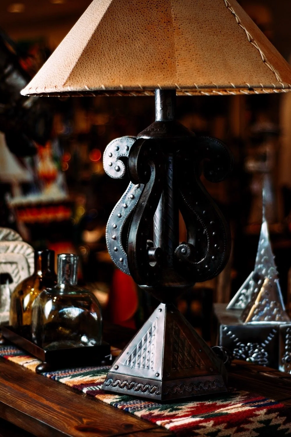 Pounded metal S shaped lamp with tan pigskin lampshade, handblown glass tequila bottles and a southwest table runner from Mexico. Visit Milagros Rústicos in Arizona today.