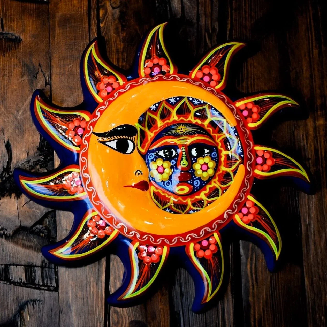 Colorful decorative sun and moon wall hanging with intricate patterns, faces, and bright colors on a wooden surface. All handmade in Mexico.