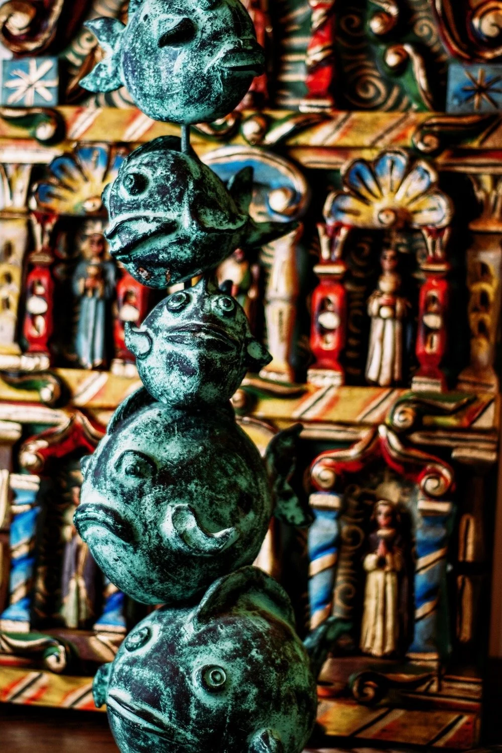 Stacked turquoise clay fish art piece in front of a gorgeous and coloful Retablo. Handmade pieces from Mexico. Visit Milagros Rústicos in Arizona today.