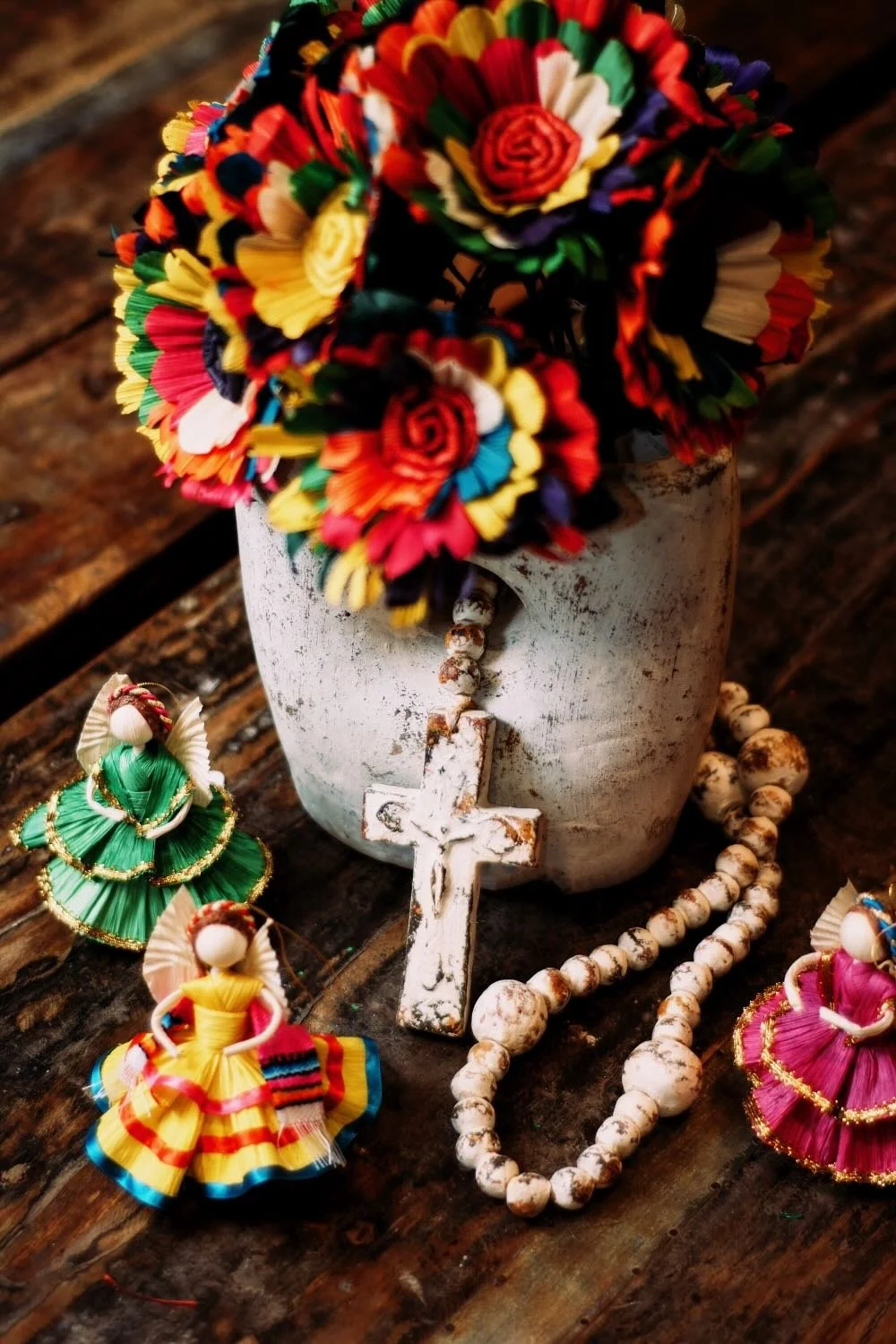 Distressed white vase filled with colorful cornhusk flowers, clay rosary beads and cornhusk Folklorico dolls from Mexico.
