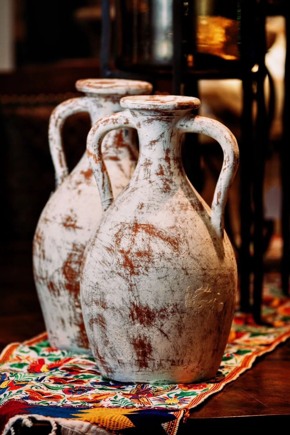 Two distressed white clay jug style vases with two handle design on a colorful embroidered table runner from Mexico.