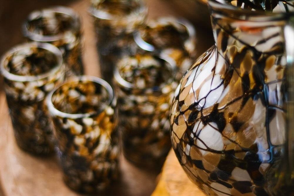 Close-up of a large mosaic handblown glass pitcher with a brown, black, and cream pattern, surrounded by several smaller matching mosaic glasses on a wooden surface. All handblown in Mexico