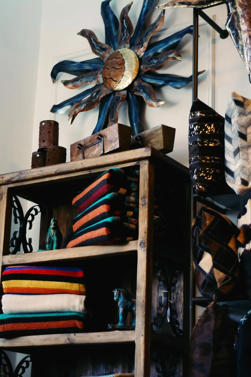 Rustic bookcase filled with Mexican blankets, clay horses, tin luminarias, vintage sugar molds below a large copper sun. 