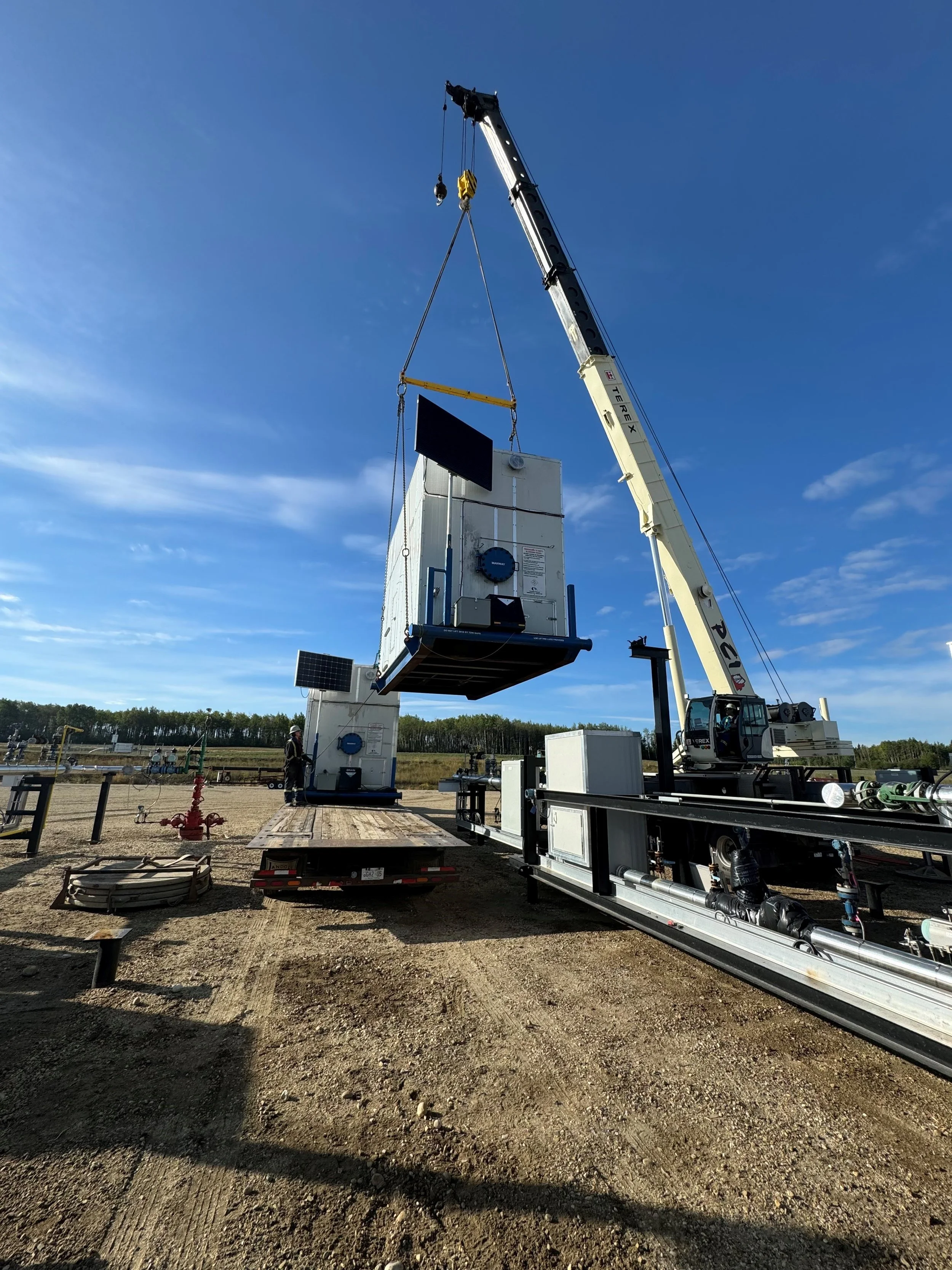 Large crane lifting and positioning a massive industrial equipment or container with solar panels attached, set against a clear blue sky on a construction or industrial site.