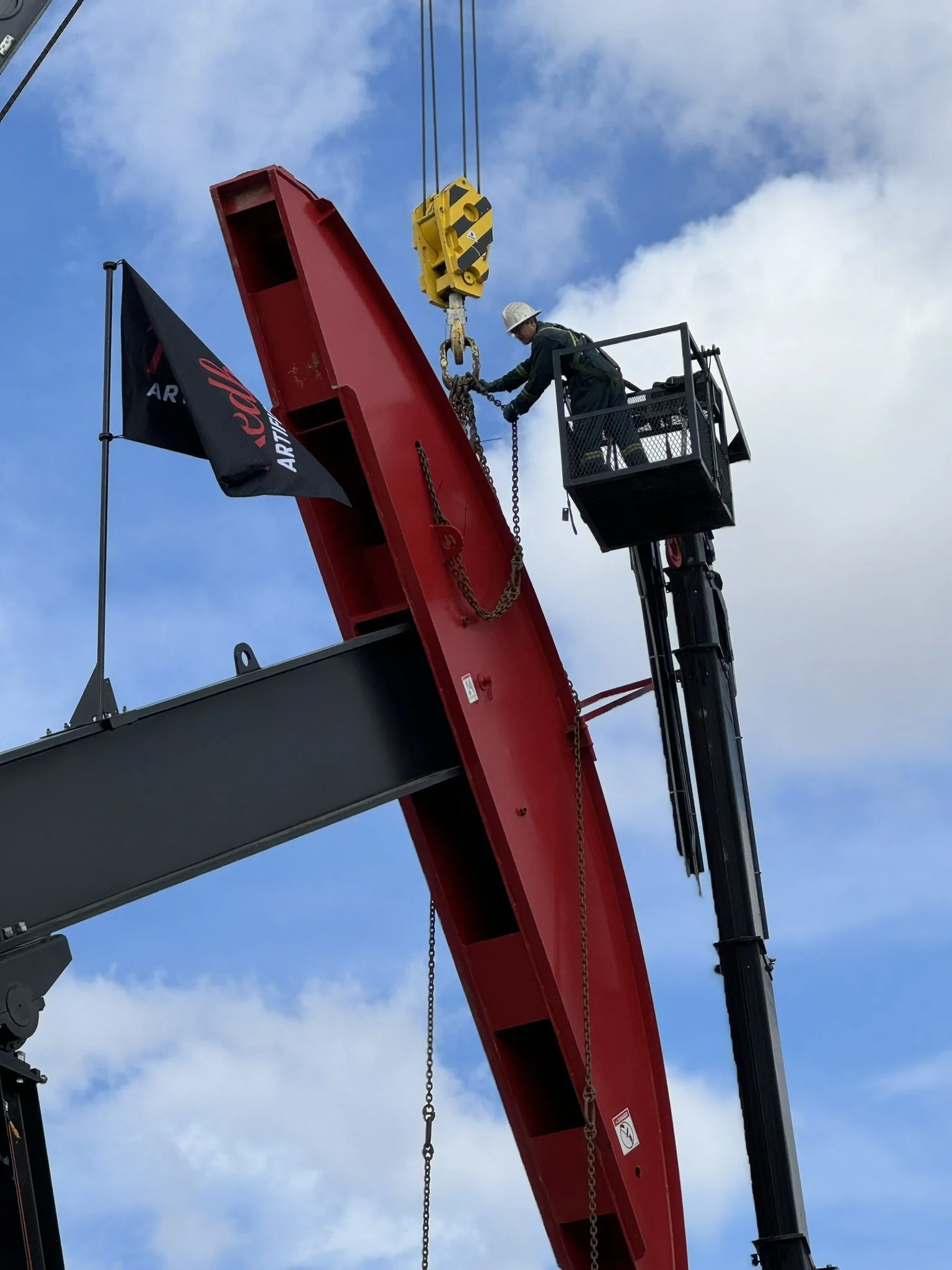 A worker in safety gear on a hydraulic lift working on a large crane component against a blue sky with clouds.