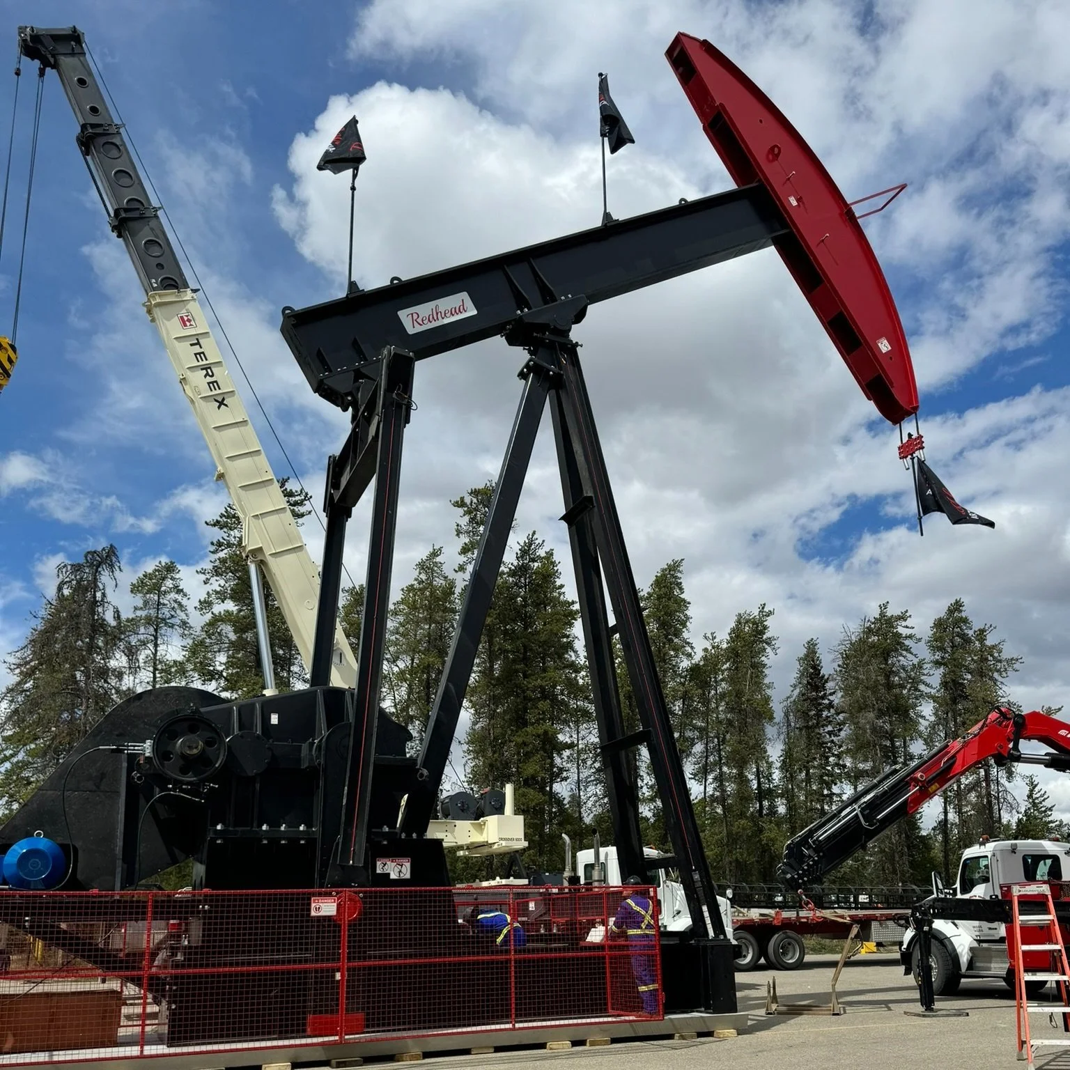 Oil pumpjack with the name 'Redhead' on it, against a partly cloudy sky and surrounded by trees.